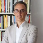 El analista y escritor Andrea Rizzi, con traje claro y gafas, delante de una estantería con libros